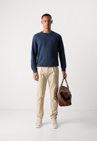 Young man wearing a navy sweatshirt and beige pants holds a brown leather duffel bag, standing against a plain white background.