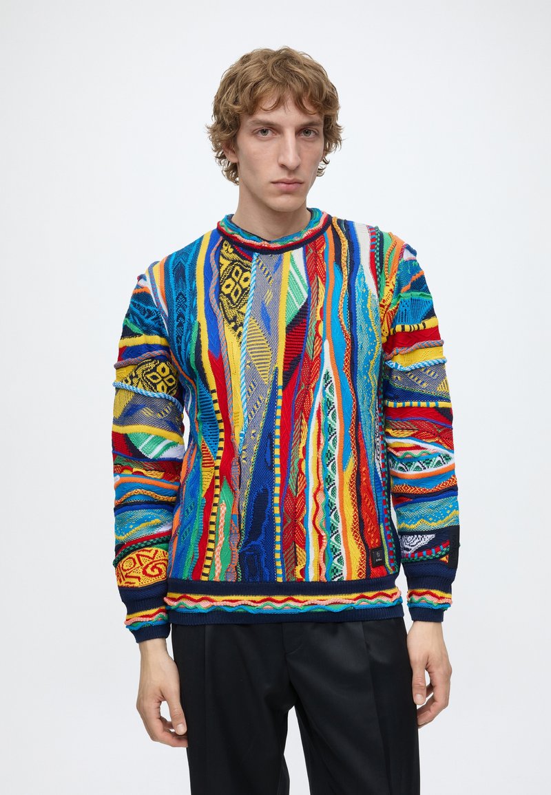 Jeune homme portant un pull coloré à motifs avec des rayures verticales et des dessins géométriques, debout devant un fond blanc uni.