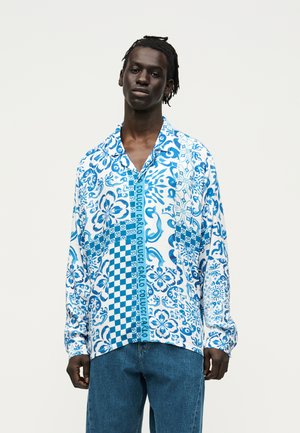 Mężczyzna ubrany w niebiesko-białą koszulę zapinaną na guziki z kwiatowym i kratkowanym wzorem oraz napisem „Carlo Colucci”, w połączeniu z niebieskimi dżinsami, stojący na białym tle.