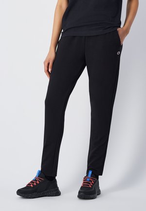 Schwarze Sporthosen aus weichem Material, mit slim-fit Schnitt, seitlichen Taschen und dezentem Logo in der Nähe des Bundes. In Kombination mit schwarzen Sneakers getragen.