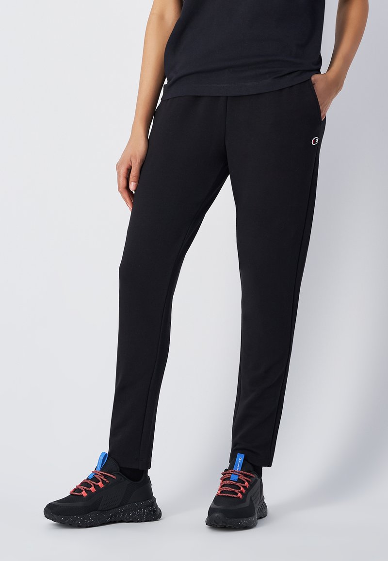 Zwarte sportbroek van zacht materiaal, met een slim fit, zijzakken en een subtiele logo nabij de tailleband. Gemaakt met zwarte sneakers.