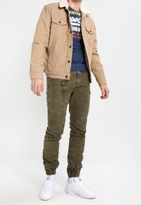 Brun denimjacka med fårskinnskrage, blå grafisk t-shirt, olivgröna joggerbyxor och vita sneakers. Avslappnad outfitkombination.