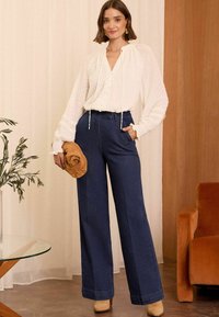 Bijela bluza s točkicama dugih rukava, V-izrezom i vezicom. Tamnoplave hlače širokih nogavica i smeđa torbica nadopunjuju izgled.