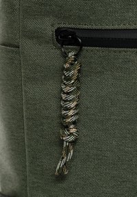 Tessuto verde texturizzato con una tasca con zip dotata di un cordino intrecciato nei colori verde, nero e marrone. Meccanismo della zip in metallo visibile.