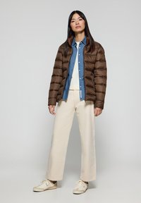 Brązowa kurtka puchowa z poziomym przeszyciem, dżinsowa koszula zapinana na guziki, szerokie spodnie w kolorze kremowym i kremowe sneakersy. Gładkie tło.