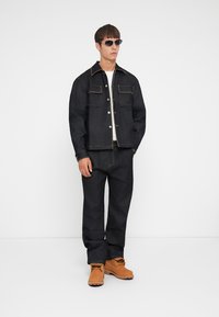 Donkerdenim jack met contrasterende stiksel, twee borstzakken, gecombineerd met bijpassende donkere spijkerbroek en bruine werklaarzen. Vlakke textuur.