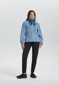 Frau steht in einer blauen Fleece-Jacke mit Reißverschluss, schwarzen Hosen und schwarzen Turnschuhen vor einem neutralen Hintergrund.