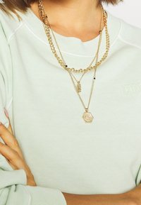 Guldpläterade halsband med olika länkar och en hexagonformad berlock, bärs över en ljusgrön sweatshirt med embossad logotyp.