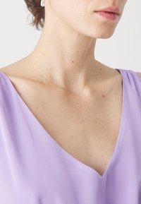 Λεβάντα αμάνικο τοπ με βαθύ V-λαιμό, κατασκευασμένο από λείο, ελαφρύ ύφασμα με διακριτική γυαλάδα. Αναδεικνύει τη κλειδώνα και τους ώμους.