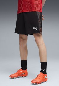 Czarne sportowe szorty o fakturowanym wzorze z białym logo Puma. W zestawie z jasnopomarańczowymi korkami piłkarskimi i czarnymi skarpetami.