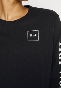 Sort langærmet t-shirt i bomuld med en hvid rektangulær "Huf."-logo på venstre bryst og hvid tekst langs ærmet.