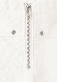 Tejido de mezclilla blanca con un cierre metálico plateado y botones de presión presenta un patrón texturizado, sugiriendo un diseño duradero y estructurado.