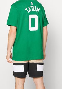 Zelené bavlněné tričko s bílým síťovaným číslem "0" a nápisem "TATUM" na zádech, doplněné černými šortkami s béžovými a bílými kapsami.