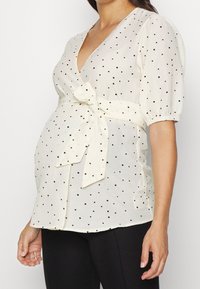 Top avvolgente per maternità in tessuto bianco con pois neri. Caratterizzato da una profonda scollatura a V, maniche a sbuffo e una cintura da annodare in vita.