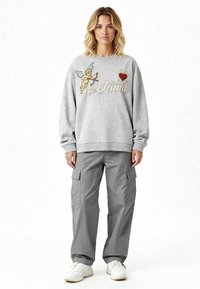 Femme portant un sweat-shirt gris avec un cupidon, un cœur et le texte « Je T’aime », assorti à un pantalon cargo gris ample et des baskets blanches, debout face à la caméra.