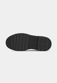 Suela de zapato de goma negra con textura acanalada, que presenta un logotipo circular y ranuras horizontales para tracción. Diseño de punta redondeada y plana.