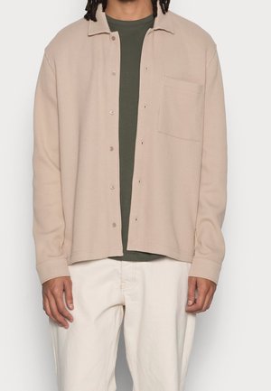 Uomo che indossa una camicia beige a bottoni con texture sopra una t-shirt verde scuro e pantaloni bianco sporco, in piedi davanti a uno sfondo semplice.