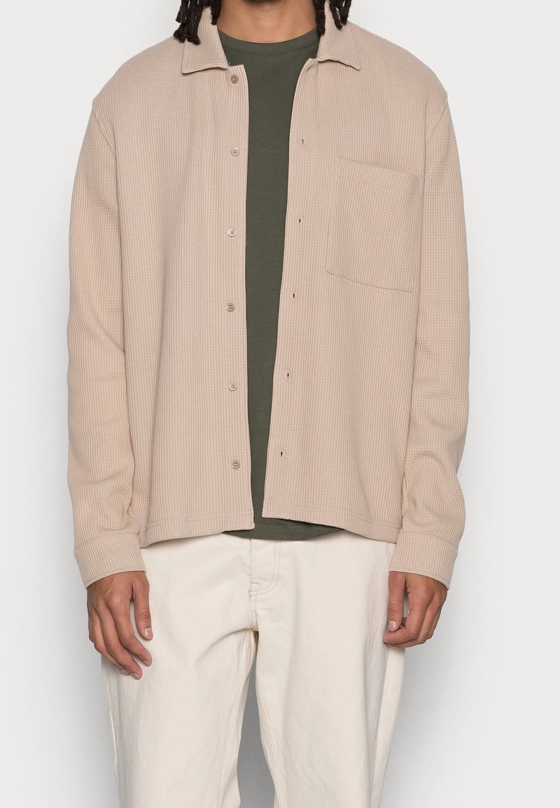 Homme portant une chemise beige à boutons texturée sur un t-shirt vert foncé et un pantalon blanc cassé, debout devant un fond uni.