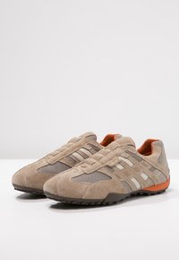 Beige och orange avslappnade sneakers med mocka- och mesh-paneler, gummisula och slip-on design placerade sida vid sida på en vit yta.