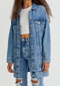 Světle modrá džínsová bunda s knoflíkovým zapínáním, dvěma náprsními kapsami a mírně oversized střihem, s ošuntělým lemem a vybledlými detaily.