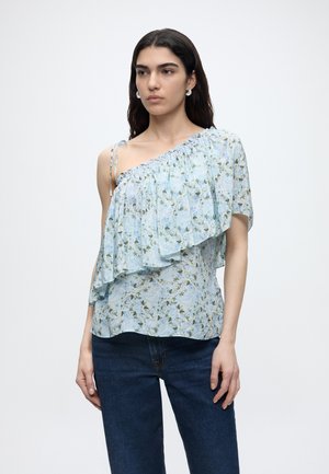 Νέα γυναίκα με μακριά σκούρα μαλλιά, φορώντας μπλε φλοράλ τοπ με ένα ώμο και βολάν και σκούρα μπλε τζιν, όρθια μπροστά σε λευκό φόντο.