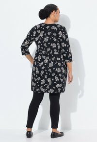 Frau mit dunklem Haar im Dutt, die eine schwarze Paisley-Tunika, schwarze Leggings und schwarze Ballerinas trägt, steht vor einem weißen Hintergrund und ist abgewandt.
