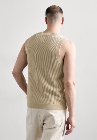 Hombre con cabello corto, vistiendo un top de punto sin mangas beige y pantalones de color claro, de pie contra un fondo claro liso, de espaldas.