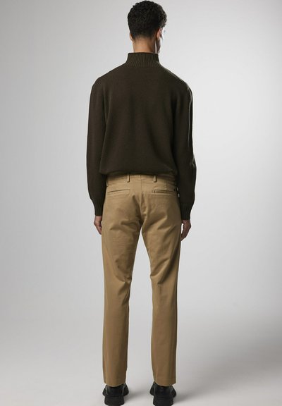 Bruine gebreide trui met hoge kraag, gecombineerd met beige broek. De broek heeft twee achterzakken en een slim fit. Model draagt zwarte schoenen.