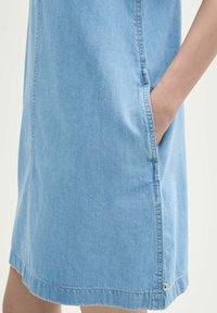 Lichtblauwe denim jurk met zijzak en zichtbare stiksels. Iets losse pasvorm, knielang met een rechte zoom en subtiele hardware-details.