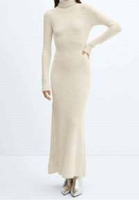 Robe à col roulé beige en texture côtelée, coupe ajustée, manches longues, et ourlet jusqu'au sol ; portée avec des escarpins pointus argentés.