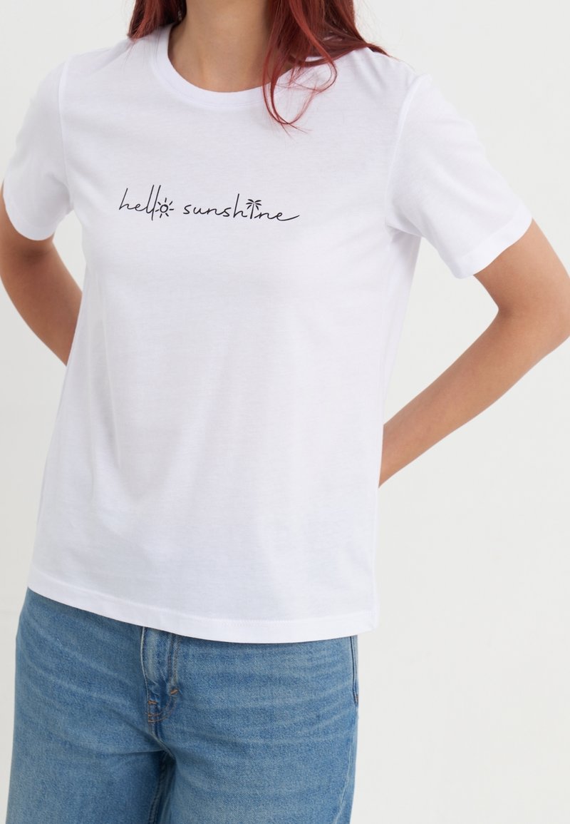 Camiseta blanca de algodón de manga corta, con texto negro escrito a mano "hello sunshine" en el frente. Combinada con denim azul claro.