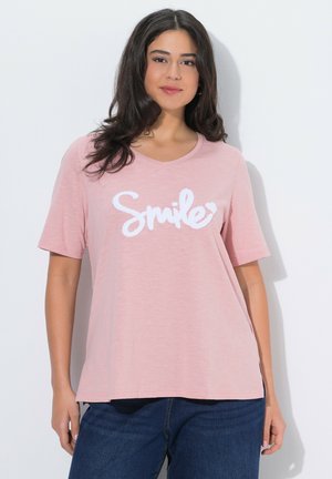 Kvinde med langt mørkt hår iført en pink T-shirt med teksten "Smile" og mørkeblå jeans, stående foran en hvid væg.