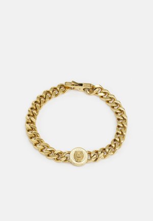 Pulsera de cadena de oro con un medallón con emblema de león y un cierre grabado con "Guess" sobre un fondo blanco liso.