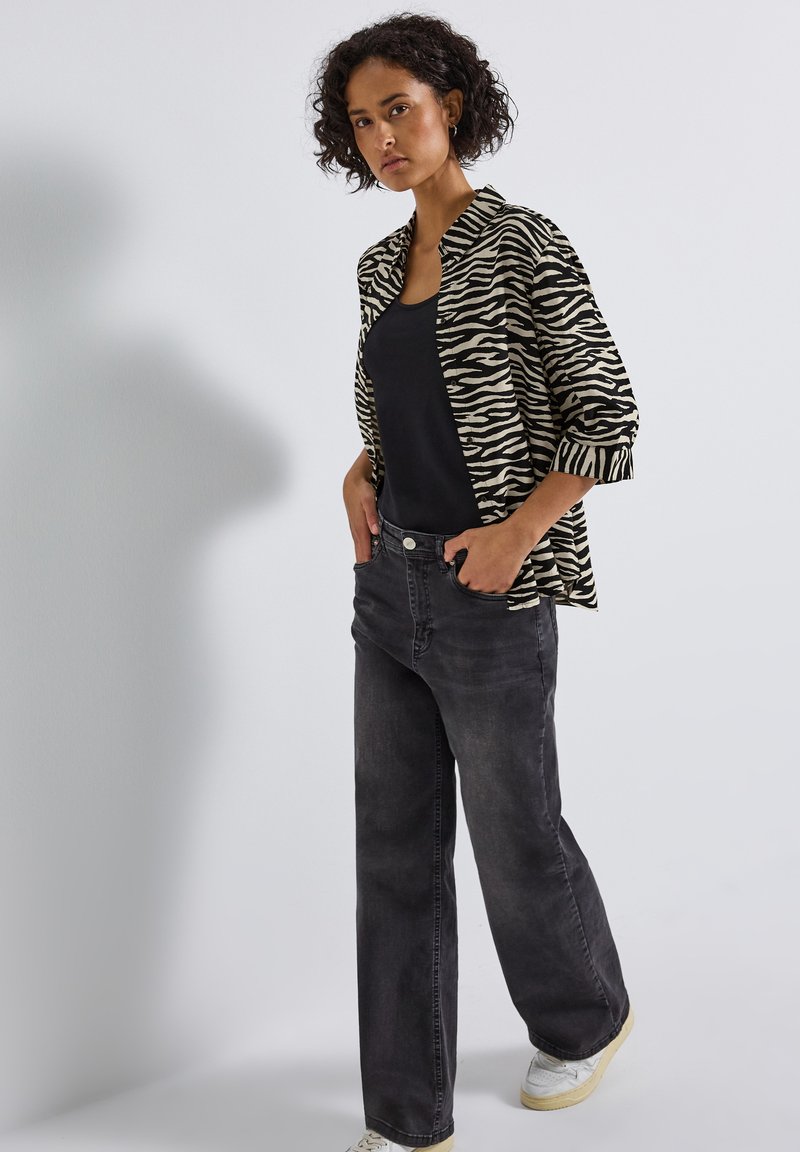 Veste imprimée zèbre beige et noir sur un haut noir, associée à un jean gris foncé et des baskets blanches, avec une coupe décontractée et des manches retroussées.