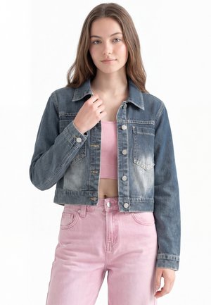 Ung kvinde med langt brunt hår iført en falmet denimjakke, pink crop top og højtaljede lyserøde jeans, står mod en hvid baggrund.