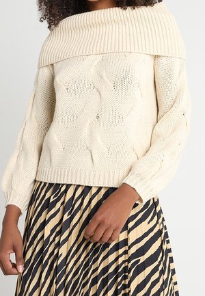 Woman wearing a cream off-shoulder cable knit sweater and a pleated beige skirt with irregular black stripes.