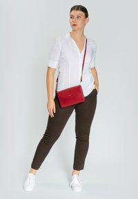 Borsa a tracolla in pelle rossa con finitura testurizzata, hardware in argento e tracolla regolabile. Il modello indossa una camicia bianca a bottoni e pantaloni marroni.