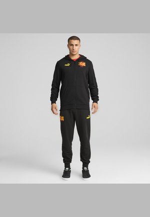 Mandlig model iført sort Puma-træningsdragt med gule og røde logoer, stående oprejst mod en ensartet lys grå baggrund.