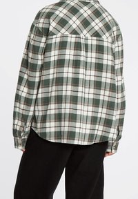Chemise en flanelle à carreaux verts et blancs avec un design à boutons, manches longues et ourlet arrondi. Texture douce avec un léger éclat.