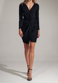 Svart klänning med lång ärm och djup V-ringning. Den har en samlad design med en draperad twist vid midjan, kompletterad med högklackade skor.