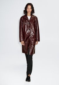 Bordeauxrode leren jas met een knoopsluiting, puntige kraag en zijzakken, gecombineerd met zwarte broek en getextureerde zwarte schoenen.