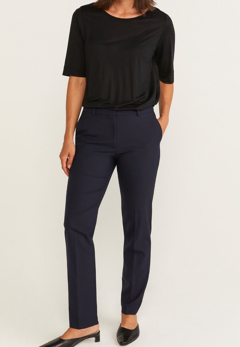 Femme portant un haut noir à manches courtes, un pantalon ajusté bleu marine et des chaussures noires à talons bas, debout avec une main dans la poche.