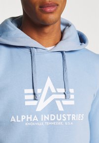 Felpa leggera blu con un logo bianco che presenta una stella astratta e delle strisce. Include cordini e una texture morbida.