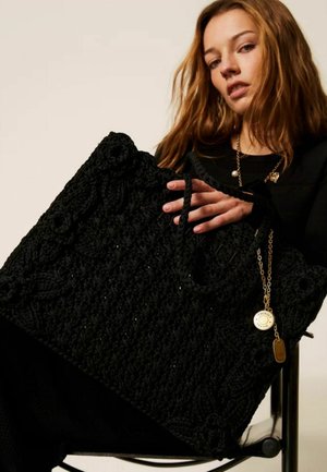 Giovane donna che tiene una grande borsa tote nera lavorata a maglia con catena dorata e ciondolo medaglione, seduta su una sedia davanti a uno sfondo neutro.