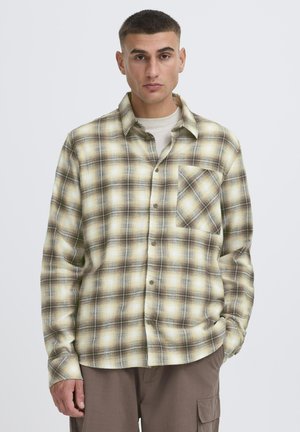 Mand iført beige og brunternet skjorte med knapper over lysebeige undertrøje og brune cargobukser, stående mod en ensfarvet baggrund.