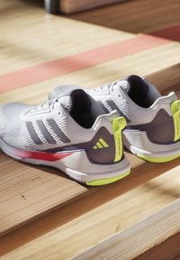 Baskets de sport grises avec tissu texturé, accents blancs et touches de vert. Présentent trois rayures noires et un détail rouge sur la semelle.