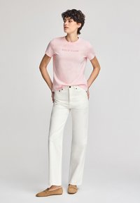 Rosa kortärmad t-shirt med "POLO CLUB" tryckt i en mörkare nyans, tillsammans med vita raka jeans och bruna mockaloafers.