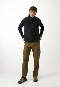 Juodas vilnonis liemenė su užtrauktuku, dviem priekiniams kišenėmis, derinamas su smėlio spalvos krovinių kelnėmis su šoninėmis kišenėmis ir juodais žygio batais.