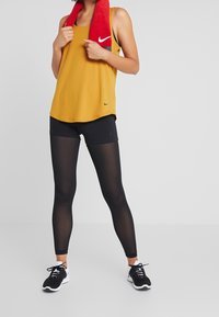 Canotta atletica gialla con accenti neri, abbinata a leggings in rete neri. Asciugamano rosso drappeggiato attorno al collo; scarpe da ginnastica nere con suole bianche.