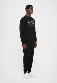 Svart hettegenser og bukser i bomullsmiks, med et gull-logo. Modellen har på seg hvite sneakers. Enkel, figurnær design med ribbet mansjetter.